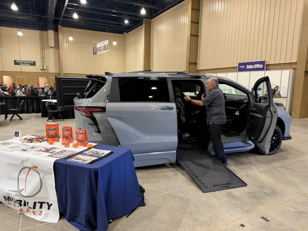 A wheelchair van specialist is standing at the open door of the Toyota Sienna wheelchair van with ramp deployed, as he is showing the interior of the van to a woman seated in a wheelchair inside the van.