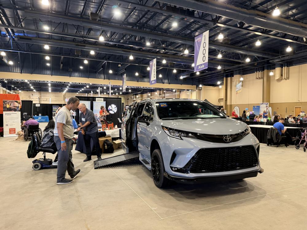 A man and woman (the woman in a wheelchair) are shown a 2026 Toyota Sienna wheelchair van by a wheelchair van specialist at the Mobility Express booth at Abilities Expo Orlando 2026.