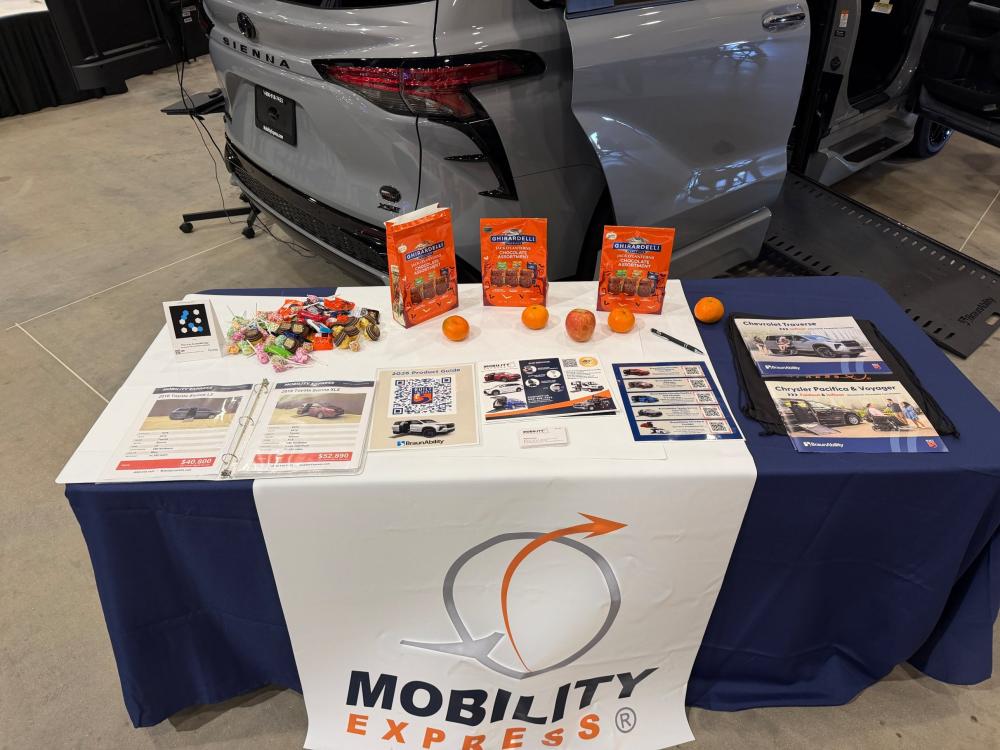 The table at the Mobility Express booth at Abilities Expo. On the table are brochures for Mobility Express, a binder with pages for wheelchair accessible vehicles in Mobility Express' inventory for guests to browse, as well as some candy and fruit for the guests, and a Mobility Express banner draped over the front of the table (white banner with black and orange text). 