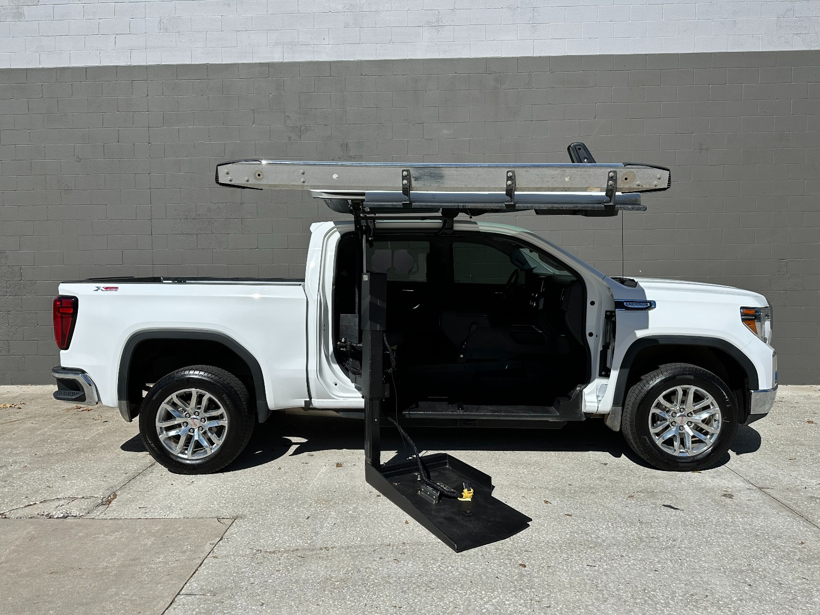 White GMC Sierra wheelchair accessible truck with lift deployed from open passenger side doors.