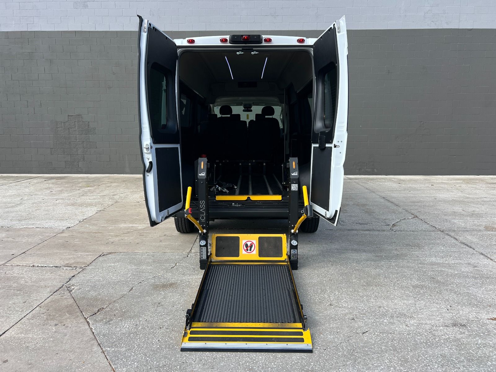 White RAM ProMaster full size wheelchair van with lift deployed from open rear barn doors. Large gray wall in the background.