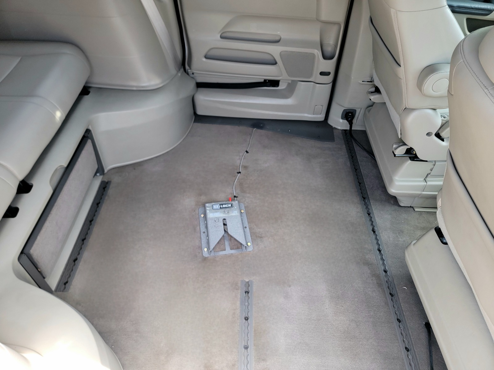 As seen from the interior of a wheelchair van, a gray square metal dock is bolted down to the middle of the floor ,which is carpeted, in the middle section of a wheelchair van.