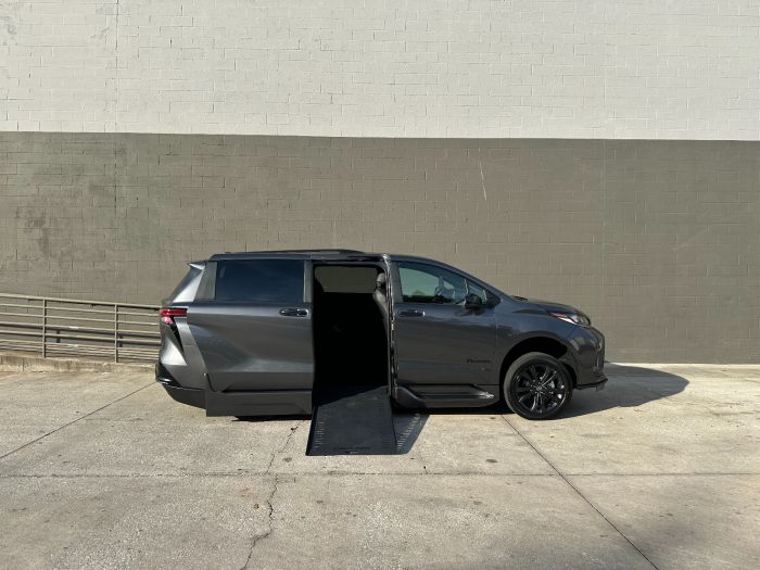 2025 BraunAbility Toyota Sienna XSE Wheelchair Van in Magentic Gray For Sale Facing the passenger side of a gray Toyota Sienna minivan. The passenger side sliding door is open, and a wheelchair ramp is deployed down to the ground. The background is a white and gray block wall.