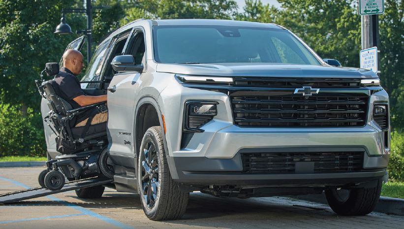 A man in a mostly black colored power wheelchair is riding it up a ramp into a Chevrolet Traverse wheelchair accessible SUV. The man and the power chair are both partially inside the vehicle.