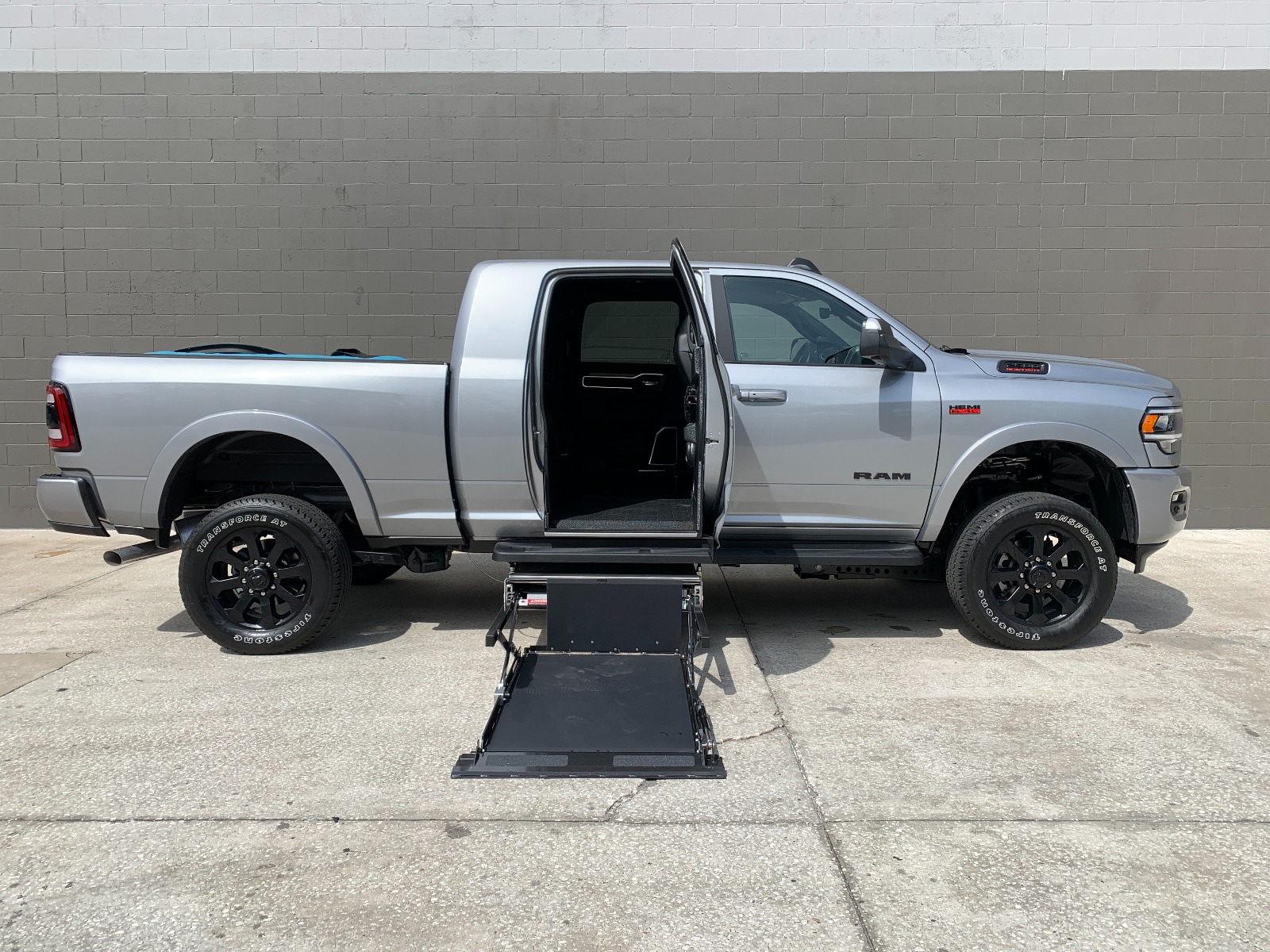 Silver RAM 2500 wheelchair accessible truck with lift deployed from open passenger side rear door.