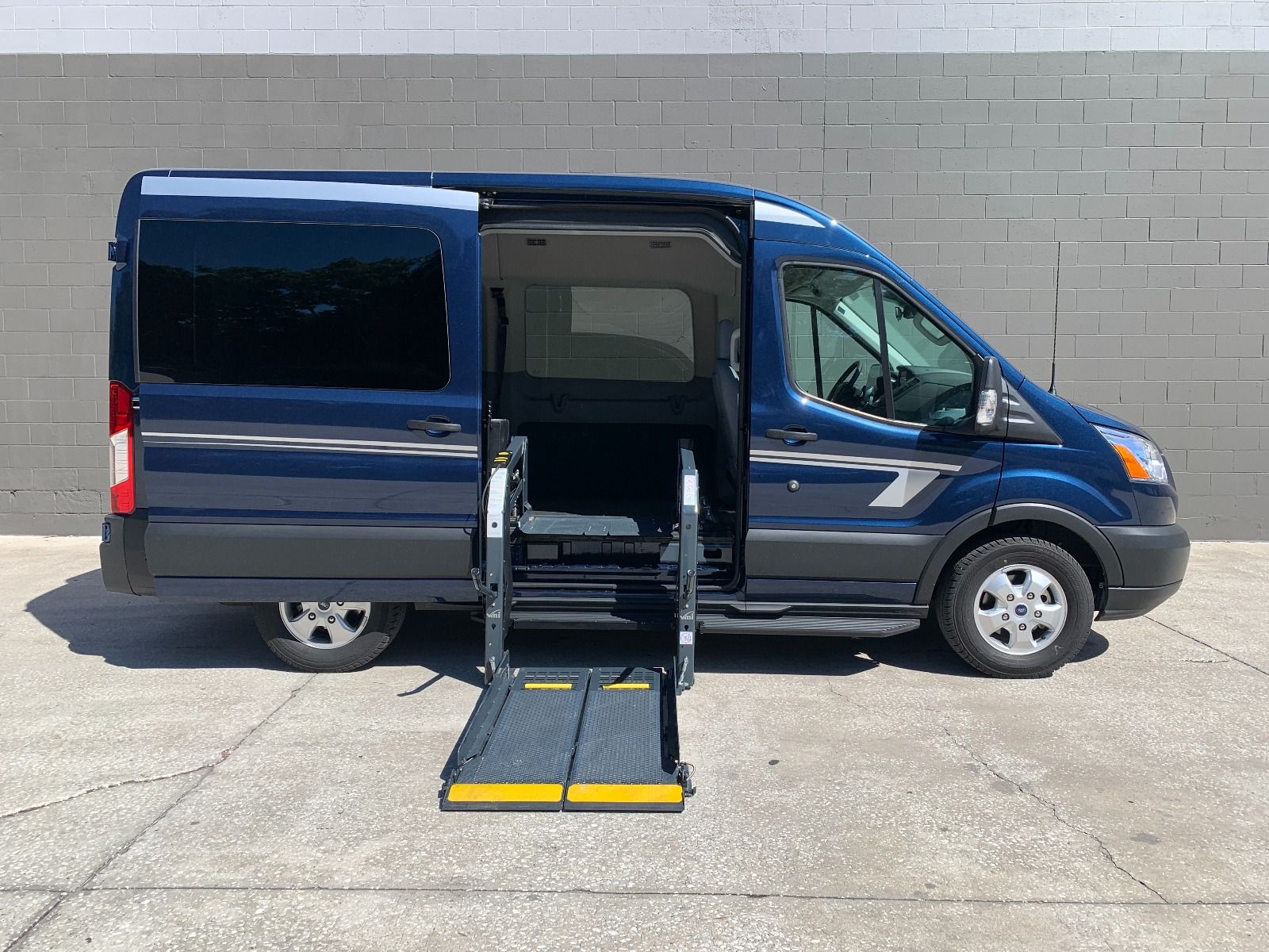 Blue Ford Transit full size wheelchair van with the wheelchair lift deployed from the open passenger side sliding door.