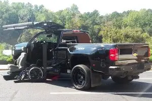 A wheelchair accessible truck with a wheelchair being loaded at the driver door, with a scenic background.